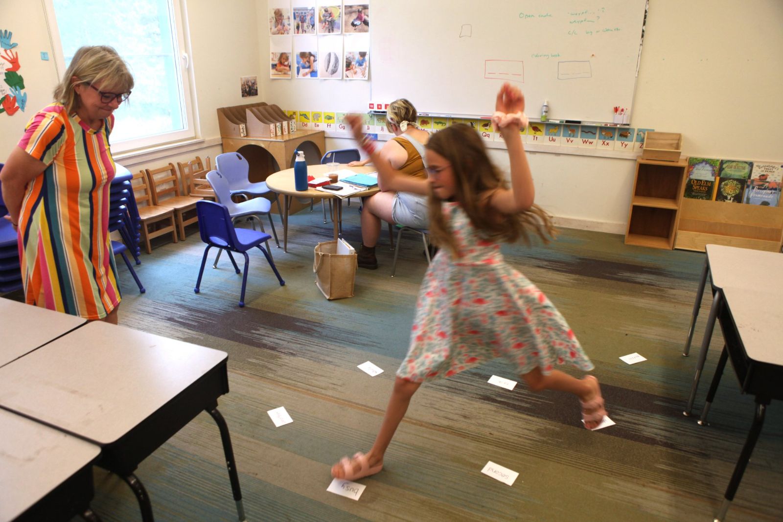A student mid-leap across word cards laid out on the floor while her tutor watches